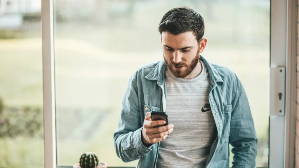 Homem de camiseta branca e jaqueta verde segurando smartphone nas mãos